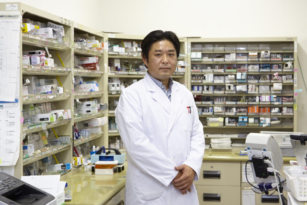介護施設の医務室。薬剤師が医薬品管理室に立つ。整理された薬品棚と医療機器が見える。